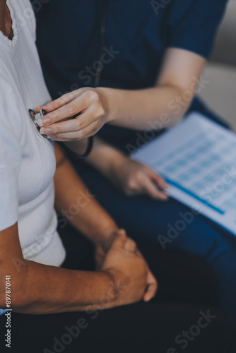 Caucasian female doctor uses stethoscope to check heart rate on elderly Asian patient on sofa.