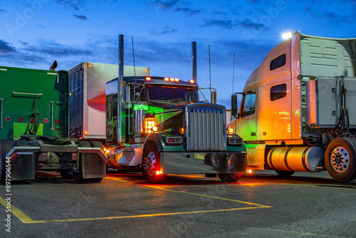 Colorful big rig semi trucks with semi trailers standing in row on the truck stop parking lot with turned on lights at night time