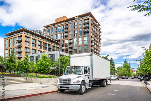 Middle duty day cab white compact bonnet rig semi truck with box trailer unloading delivered to customer apartments goods standing on the urban city street at cloudy day