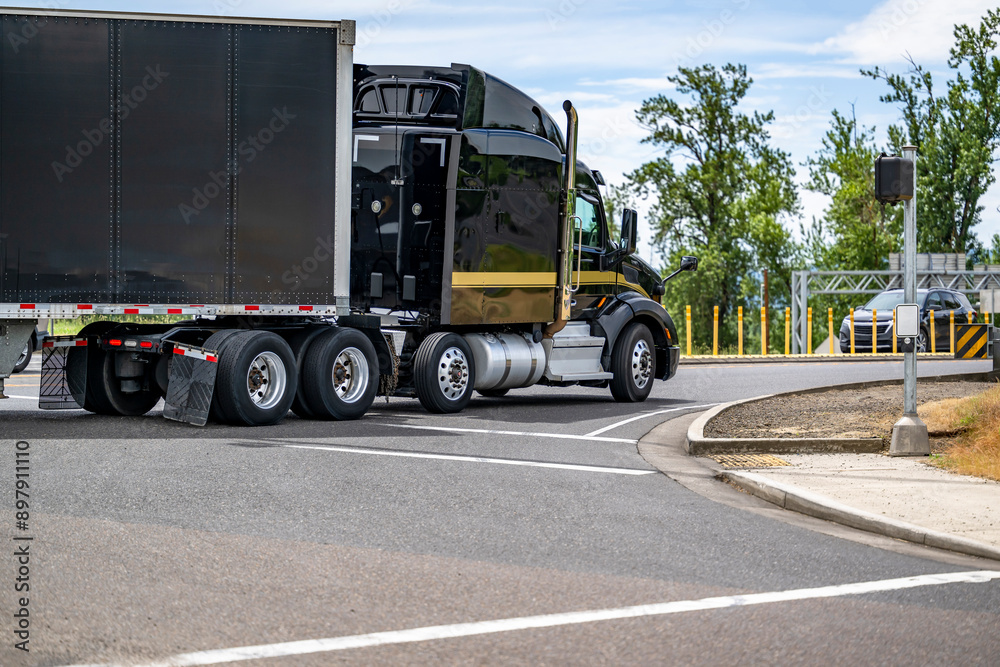 Stylish black big rig semi truck with high roof transporting cargo in tented semi trailer driving on the round road intersection Stylish black big rig semi truck with high roof transporting cargo in tented semi trailer driving on the round road intersection