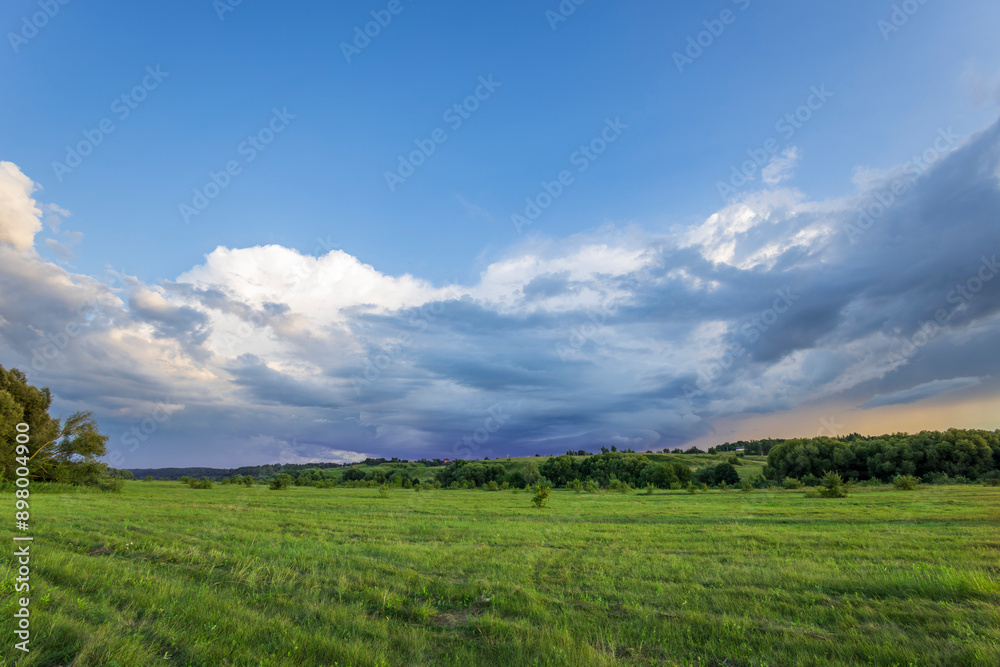 A wide expanse of green meadow stretches out beneath a dramatic sky filled with billowing white and grey clouds, hinting at the possibility of a coming storm. A wide expanse of green meadow stretches out beneath a dramatic sky filled with billowing white and grey clouds, hinting at the possibility of a coming storm.
