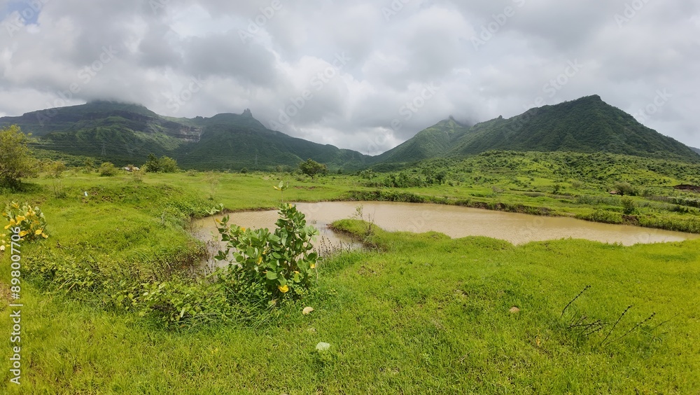 Scenic View of Sahyadri Hills with Pond, Lush Green Meadow, and Cloudy Sky during Monsoon Season Scenic View of Sahyadri Hills with Pond, Lush Green Meadow, and Cloudy Sky during Monsoon Season
