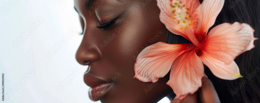 Fototapeta Close-up of a beautiful black woman with a hibiscus flower in her hair on a white background, the concept of skin care and beauty.