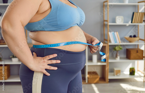 Crop shot overweight, obese woman in sports bra holding measuring tape around fat belly to check how much weight she has gained and how many centimeters she has added in her waist. Fat body concept