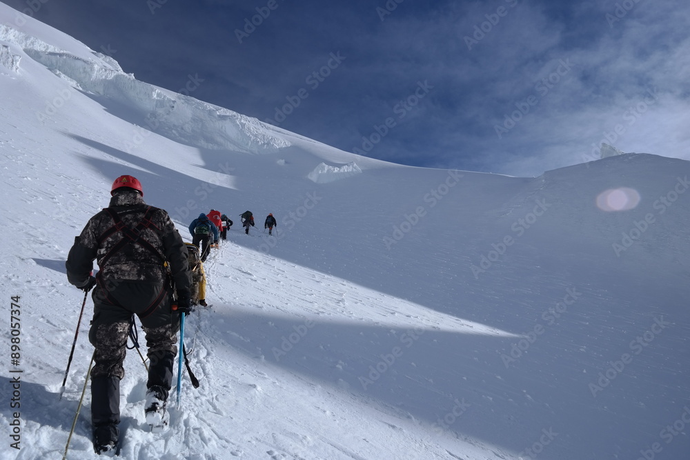 Experienced mountaineers ascend steep snowy slope on a majestic mountain, linked by safety ropes through powdery snow. Teamwork, endurance, and determination in high-altitude climb. Experienced mountaineers ascend steep snowy slope on a majestic mountain, linked by safety ropes through powdery snow. Teamwork, endurance, and determination in high-altitude climb.