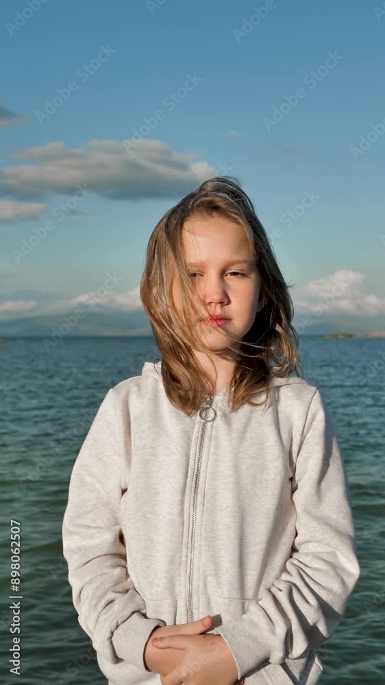 Vertical portrait of a teenage girl looking at the frame on the seashore at sunset. The child of 9 years old looks and smiles sweetly, squinting a little from the sun. The wind blows the girl's hair Vertical portrait of a teenage girl looking at the frame on the seashore at sunset. The child of 9 years old looks and smiles sweetly, squinting a little from the sun. The wind blows the girl's hair