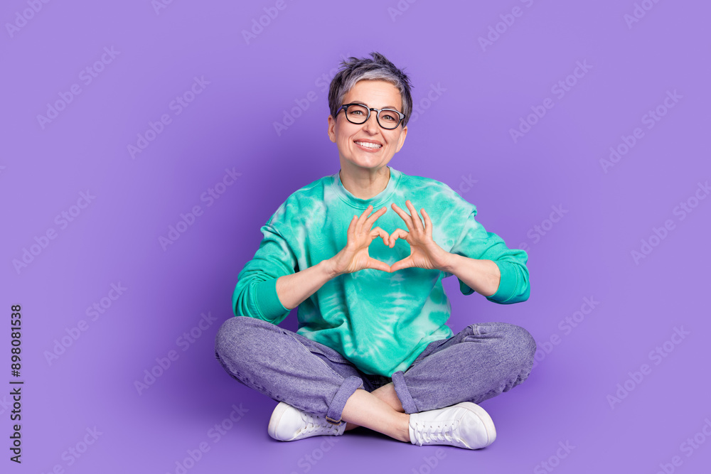 Full body photo of attractive senior woman sit floor show heart symbol dressed stylish green clothes isolated on purple color background Full body photo of attractive senior woman sit floor show heart symbol dressed stylish green clothes isolated on purple color background