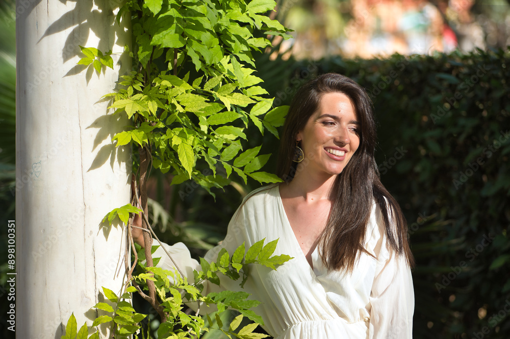 Young woman, natural, with brown hair and white dress, smiling and relaxed leaning on a marble column with vegetation. Concept naturalness, simplicity, relax, smile. Young woman, natural, with brown hair and white dress, smiling and relaxed leaning on a marble column with vegetation. Concept naturalness, simplicity, relax, smile.