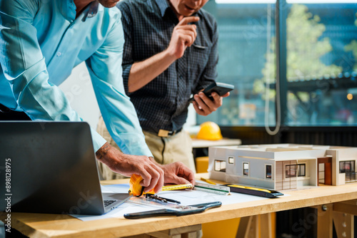 A Caucasian male engineer and an Italian male building contractor are planning and talking at a desk, reviewing house plans and a model of a house with wooden textures for renovation.