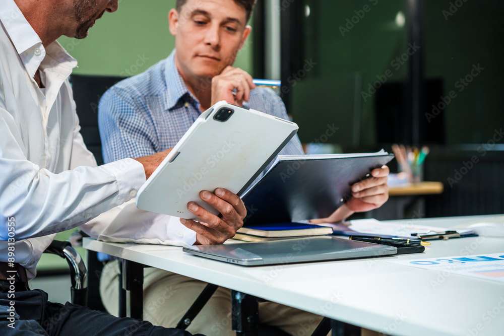 An Italian male and a Caucasian male sit at business desk in office meeting room, discussing financial terms, investment strategies, managing funds with computer monitor displaying relevant data. An Italian male and a Caucasian male sit at business desk in office meeting room, discussing financial terms, investment strategies, managing funds with computer monitor displaying relevant data.