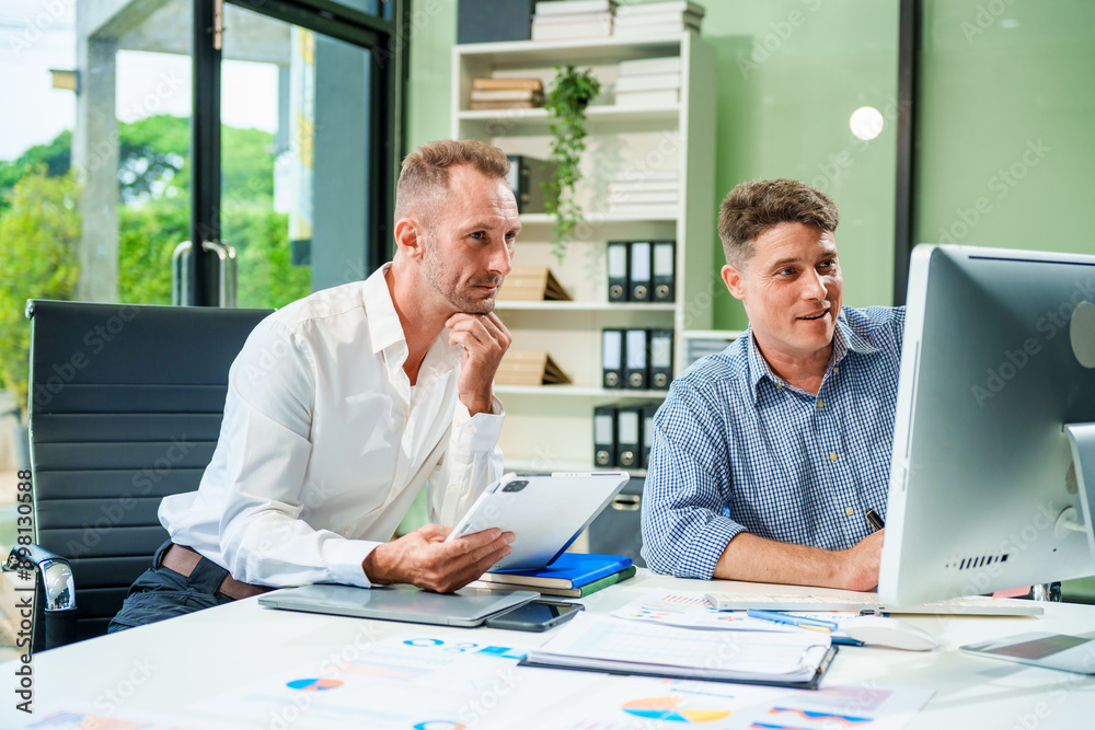 An Italian male and a Caucasian male sit at business desk in office meeting room, discussing financial terms, investment strategies, managing funds with computer monitor displaying relevant data. An Italian male and a Caucasian male sit at business desk in office meeting room, discussing financial terms, investment strategies, managing funds with computer monitor displaying relevant data.