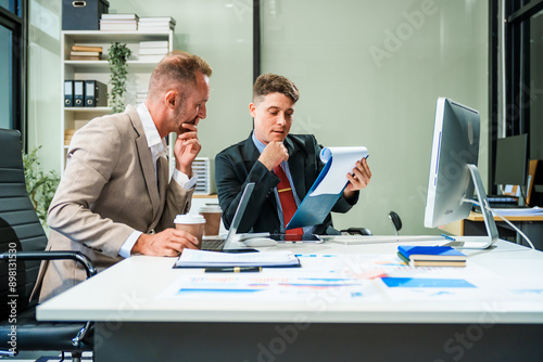 Foto An Italian male and a Caucasian male, both in formal suits, sit at a business desk in an office meeting room with a desktop monitor and laptop, discussing business finance and investment strategies