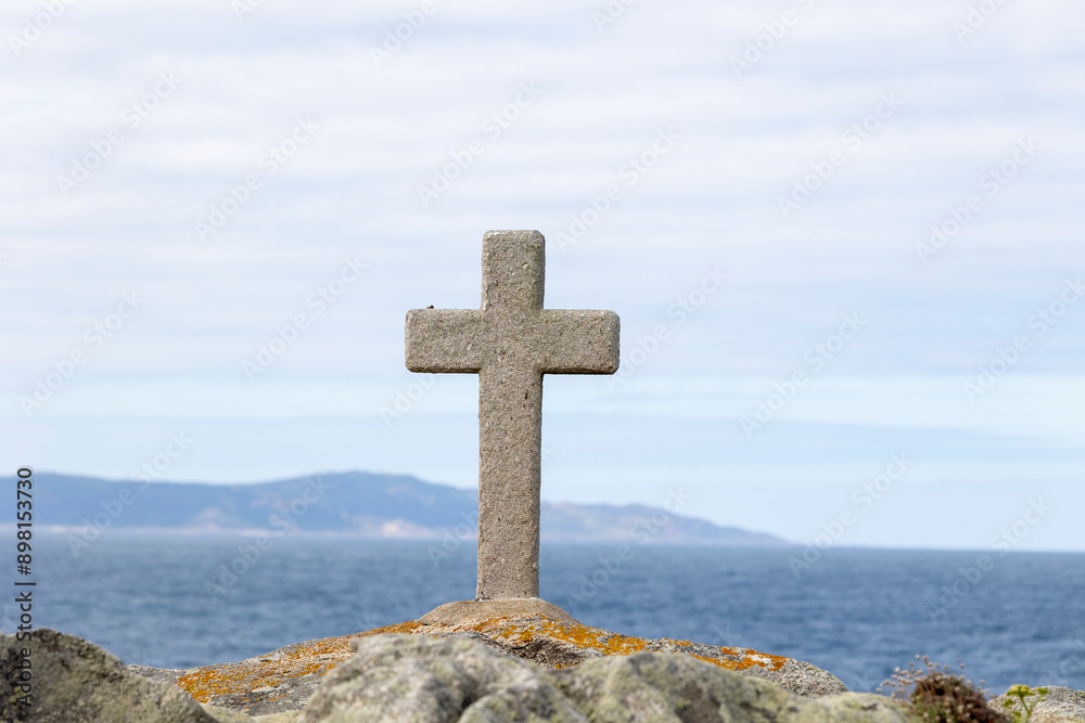 A white cross stands on rocky terrain by the seashore, with waves gently lapping and mountains faintly visible in the distance, symbolizing tranquility and solitude. A white cross stands on rocky terrain by the seashore, with waves gently lapping and mountains faintly visible in the distance, symbolizing tranquility and solitude.