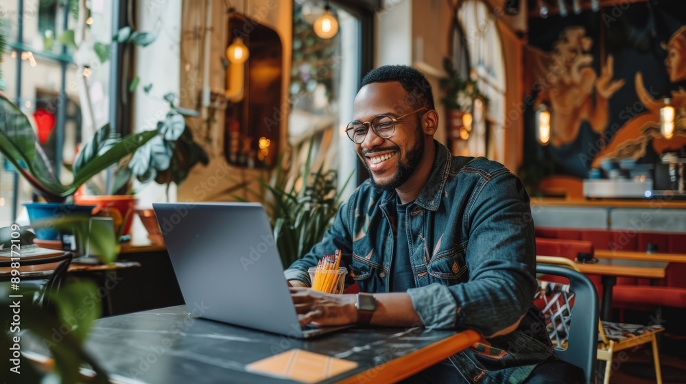 A happy man wearing glasses is using a laptop at a trendy, well-decorated cafe, suggesting a modern and upbeat work atmosphere filled with light and plants. A happy man wearing glasses is using a laptop at a trendy, well-decorated cafe, suggesting a modern and upbeat work atmosphere filled with light and plants.