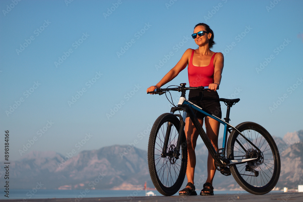 Fototapeta premium outdoor pursuit. healthy lifestyle concept. a woman with a sports bike in glasses on the beach against the background of the sky and mountains