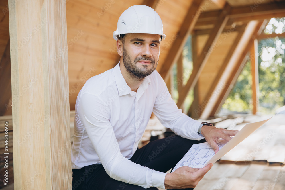 Architect wooden buildings. Man stands under frame wooden house. Man architect studies documentation or blueprints. Guy in protective helmet at construction site. Architect under timber framed house Architect wooden buildings. Man stands under frame wooden house. Man architect studies documentation or blueprints. Guy in protective helmet at construction site. Architect under timber framed house