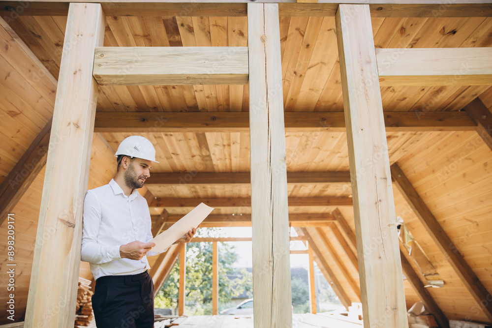 Architect wooden buildings. Man stands under frame wooden house. Man architect studies documentation or blueprints. Guy in protective helmet at construction site. Architect under timber framed house Architect wooden buildings. Man stands under frame wooden house. Man architect studies documentation or blueprints. Guy in protective helmet at construction site. Architect under timber framed house