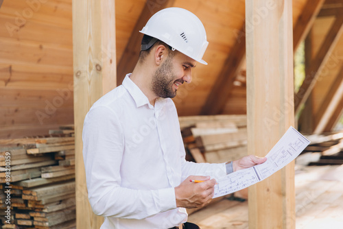Construction engineer in white helmet. Developer with construction documentation on the background of wooden building under construction. Portrait of smiling architect near eco friendly house. © anatoliycherkas