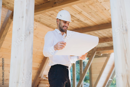 Construction engineer in white helmet. Developer with construction documentation on the background of wooden modular building under construction. Portrait of smiling architect near eco friendly house. © anatoliycherkas