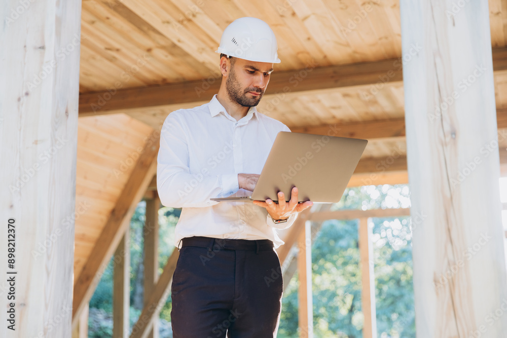 Man builder. Foreman with laptop. Contractor for construction company. Architect in unfinished building. Builder in wooden modular house under construction. Man engineer from construction team Man builder. Foreman with laptop. Contractor for construction company. Architect in unfinished building. Builder in wooden modular house under construction. Man engineer from construction team