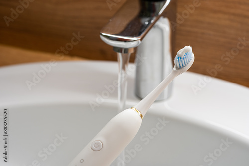 Upright electric toothbrush on sink edge, faucet background.