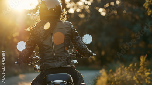 A woman on a motorcycle rides along a beautiful forest road against the backdrop of sunset, rear view. A female motorcyclist with flying hair in a leather jacket rides a motorcycle.