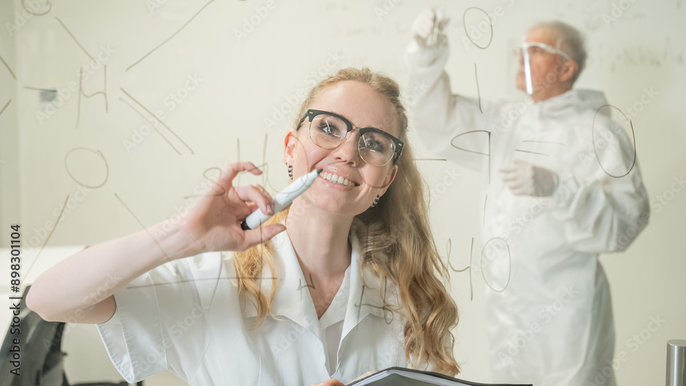 A woman chemist writes a formula on glass. An elderly Caucasian man in a protective suit is doing tests. A woman chemist writes a formula on glass. An elderly Caucasian man in a protective suit is doing tests.