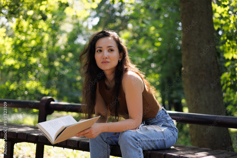 Naklejka premium Brunette asian woman holding book while sitting on bench in summer park