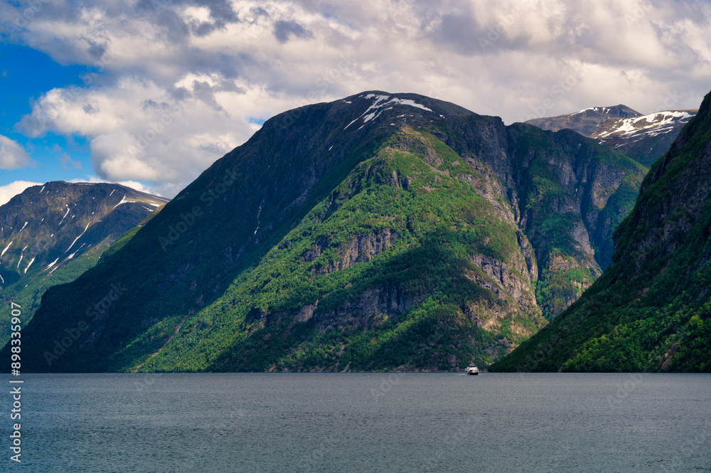 The beautiful Geiranger fjord, in Norway. A small ferry boat can be seen, dwarfed by the huge mountains, moving towards the town of Hellesylt The beautiful Geiranger fjord, in Norway. A small ferry boat can be seen, dwarfed by the huge mountains, moving towards the town of Hellesylt