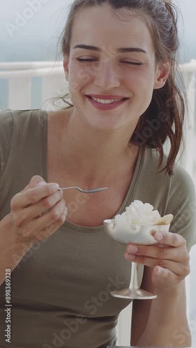 Portrait of a happy young woman, eating and enjoying a cup of gelato with whipped cream, smiling and looking at the camera. Attractive girl at terrace of cafe in sunny summer day. Vertical video
