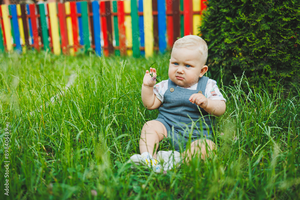 A little boy one year old sits in the green grass on the street. Summer clothes for babies. A little boy one year old sits in the green grass on the street. Summer clothes for babies.