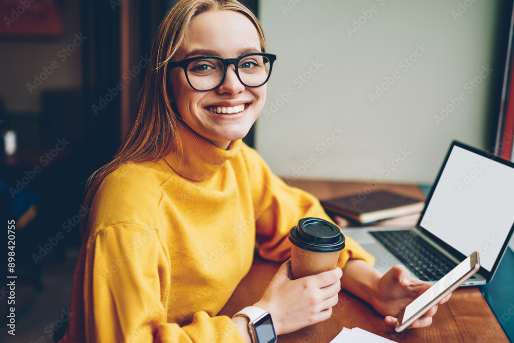 Portrait of cheerful hipster girl sitting at table with modern laptop computer with blank screen area for internet advertising while holding smartphone gadget and looking at camera during coffee break Portrait of cheerful hipster girl sitting at table with modern laptop computer with blank screen area for internet advertising while holding smartphone gadget and looking at camera during coffee break