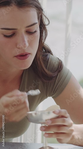 Portrait of a happy young woman, eating and enjoying a cup of gelato with whipped cream, smiling and looking at the camera. Attractive girl at terrace of cafe in sunny summer day. Vertical video