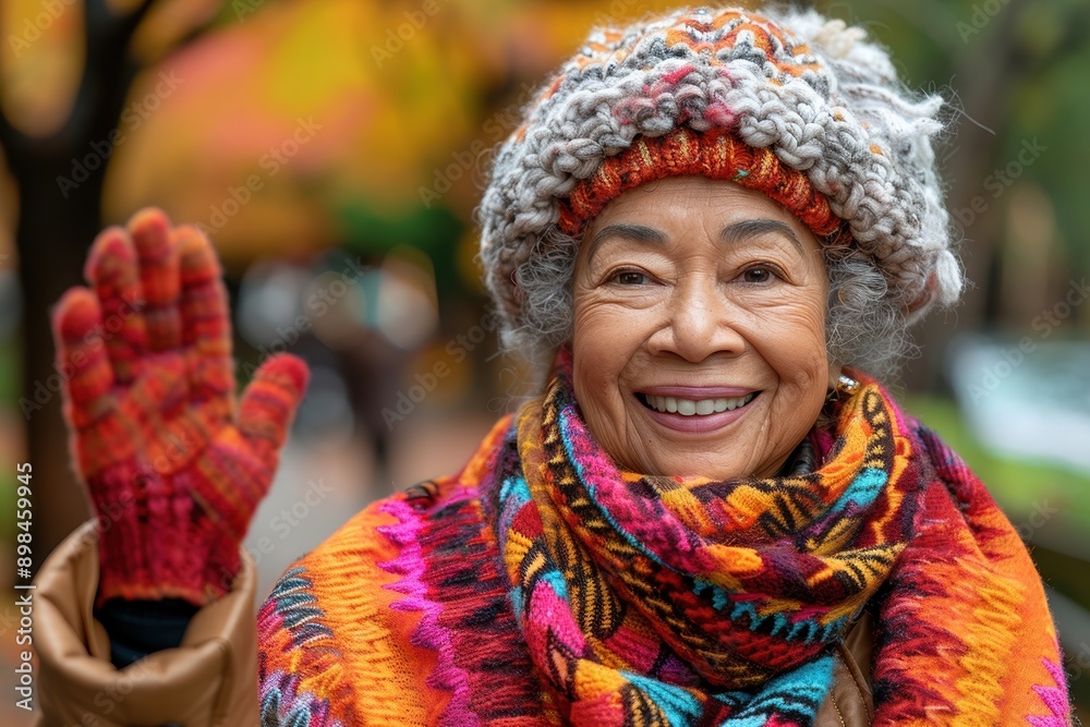 Elderly woman with a cheerful smile, wearing colorful winter wear including knit hat, scarf, and gloves, greeting while standing in a beautiful fall landscape Elderly woman with a cheerful smile, wearing colorful winter wear including knit hat, scarf, and gloves, greeting while standing in a beautiful fall landscape