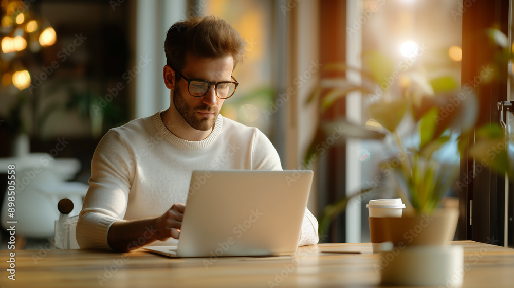 A man in glasses working intently on his laptop in a cozy cafe, surrounded by plants and warm lighting. A man in glasses working intently on his laptop in a cozy cafe, surrounded by plants and warm lighting.