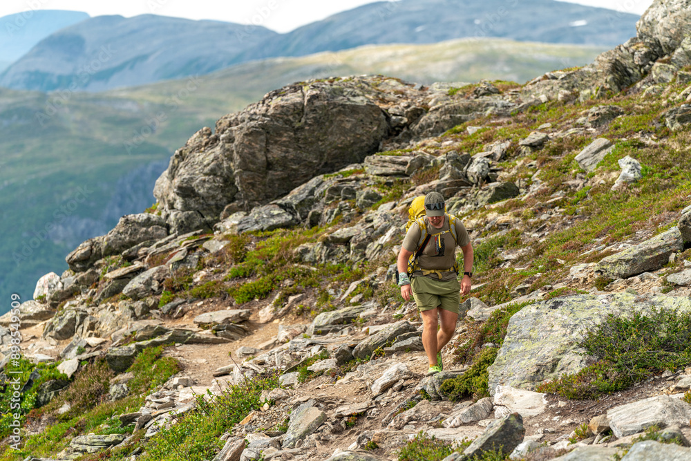 male backpacking hiker with yellow backpack climbing Romsdalseggen trail in Andalsnes Norway in summer with rocky mountains in the background male backpacking hiker with yellow backpack climbing Romsdalseggen trail in Andalsnes Norway in summer with rocky mountains in the background