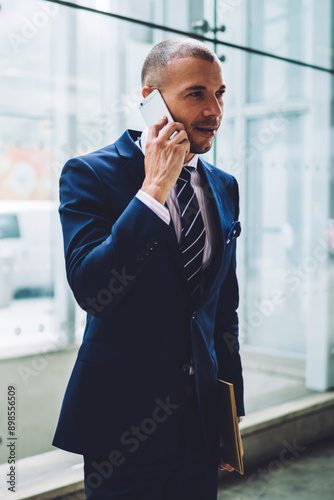 Professional male trader dressed in stylish elegant suit calling on smartphone device.Successful young businessman communicating on telephone device standing indorr in office building photo