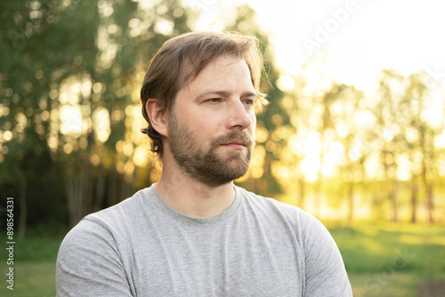 Portrait of handsome young man in gray t-shirt outdoors. Male with long hair and beard feel happy, calm and serious on Green nature sunset background. Man looking away into distance. Diversity People © Marina Demidiuk