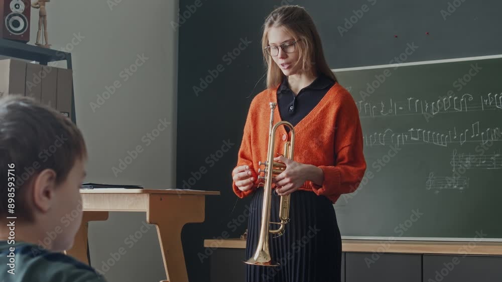 Young female teacher holding trumpet and explaining how to play it while standing in front of primary school students in music class