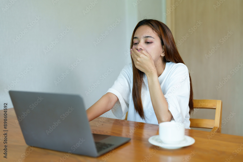 Asian woman in white shirt yawning while working on laptop at wooden table. Background includes a coffee cup. Captures tired, unmotivated moment of remote work.