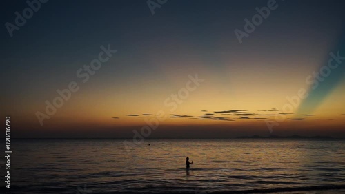 Silhouette of woman stands in the peaceful sea with beautiful twilight sky in background. Golden sky with the horizontal line of seascape. Explosion of sunlight in the evening sky during the sunset