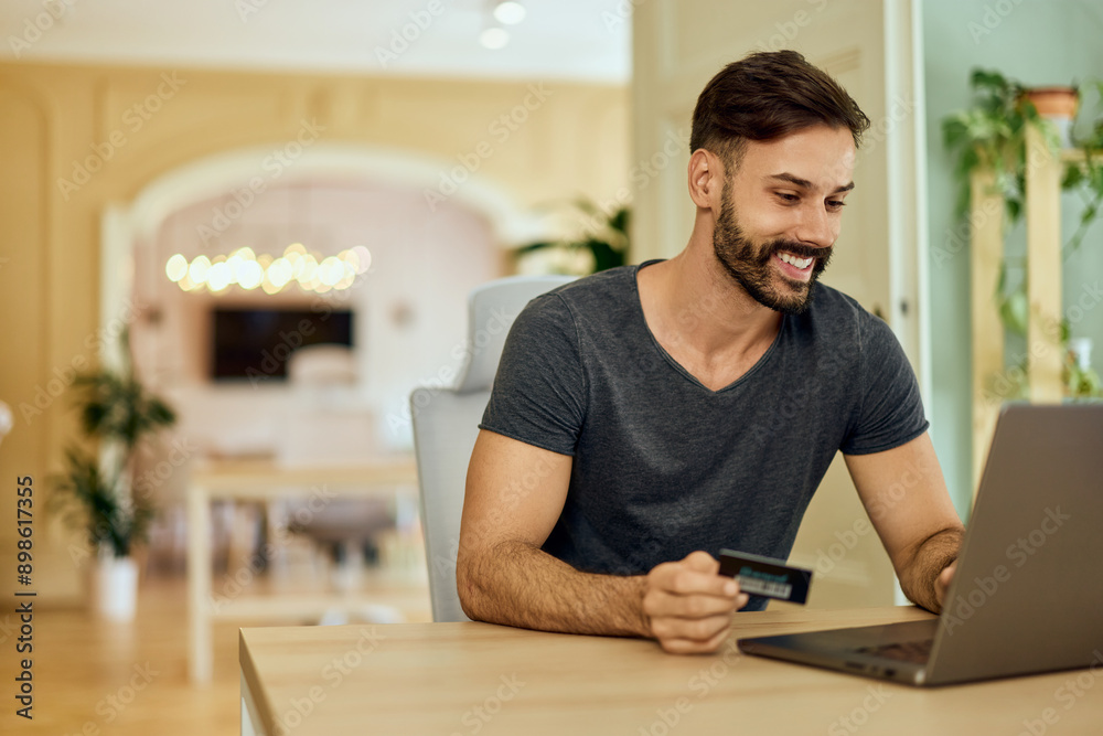 A smiling freelance man, using a credit card and typing something on the laptop.