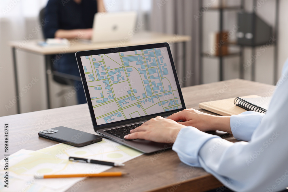 Cartographer working with cadastral map on laptop at wooden table in office, closeup Cartographer working with cadastral map on laptop at wooden table in office, closeup