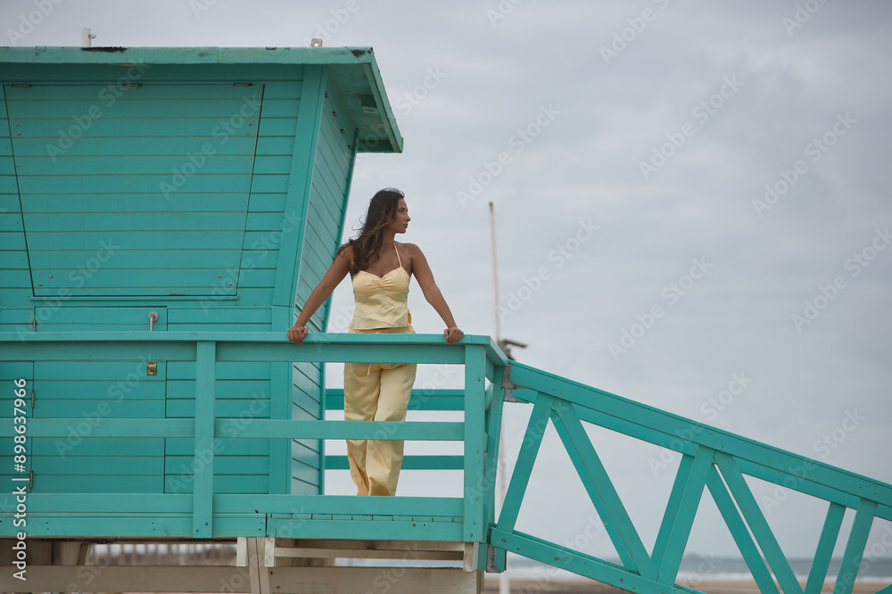 Beautiful young woman with green eyes in a yellow silk suit and pink heels, looking at the sea, leaning on a railing of a green lifeguard booth. Concept beauty, fashion, beach, coast, ocean. Beautiful young woman with green eyes in a yellow silk suit and pink heels, looking at the sea, leaning on a railing of a green lifeguard booth. Concept beauty, fashion, beach, coast, ocean.