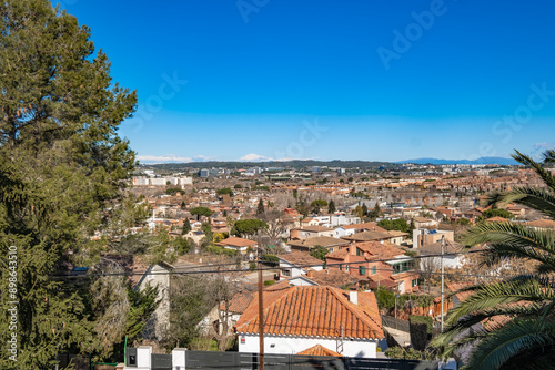 The image captures a scenic view of a cityscape from an elevated point, showcasing red brick rooftops, green foliage, and a clear blue sky stretching into the horizon Sant Cugat in Spain