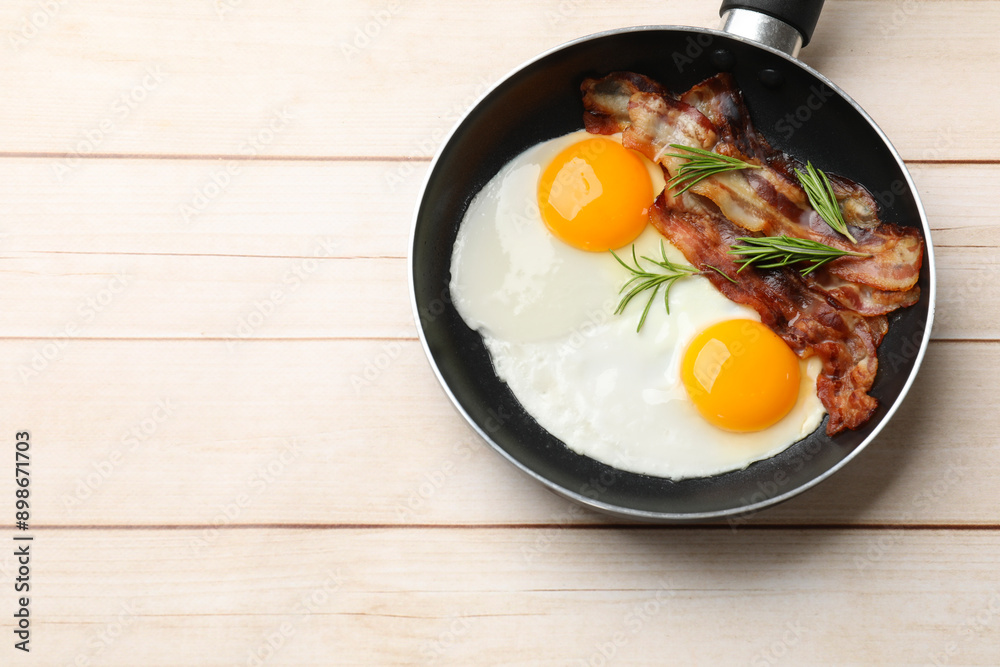 Tasty bacon, eggs and rosemary in frying pan on light wooden table, top view. Space for text Tasty bacon, eggs and rosemary in frying pan on light wooden table, top view. Space for text