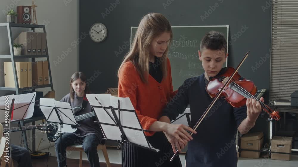 Young female intern teaching teen boy to play violin and holding bow together with him, then helping him with notes, children relaxing in background