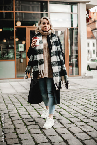 A woman wearing a plaid scarf and a sweater is walking down a street holding a coffee cup