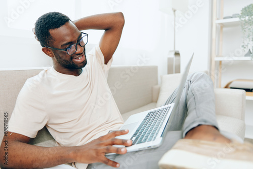 Smiling African American Freelancer Working on Laptop in Modern Home Office, Typing and Communicating Online This image captures a young, happy African American man sitting on a sofa, fully absorbed