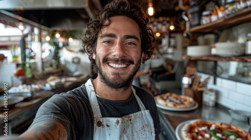 Happy young man wearing an apron, smiling for a selfie, standing in front of a counter with homemade pizza in a modern cafe kitchen.